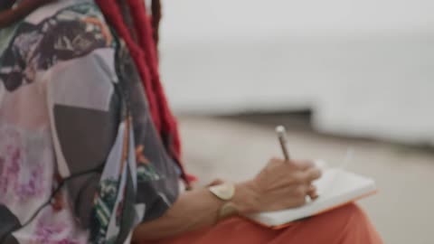 An artistic woman with red dreadlocks writes in her journal while sitting peacefully by the water