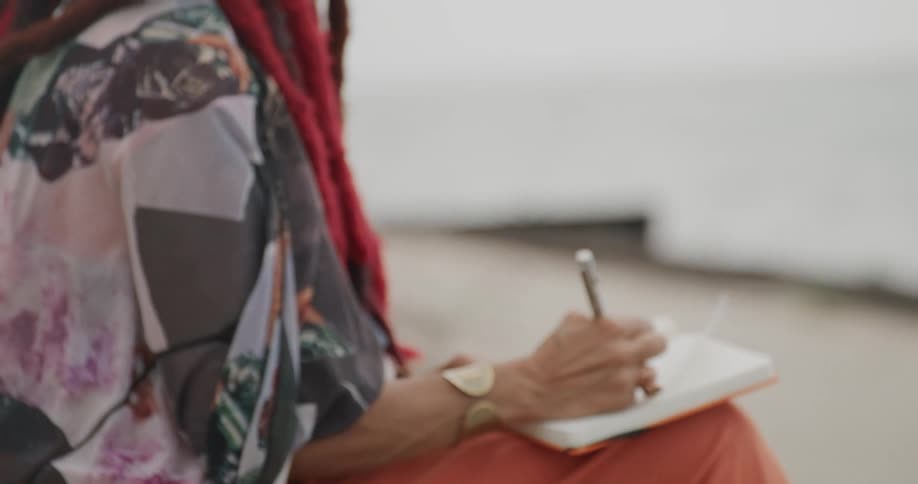 An artistic woman with red dreadlocks writes in her journal while sitting peacefully by the water