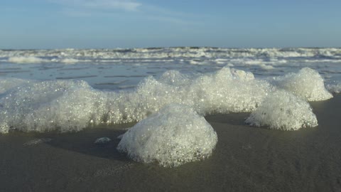 A low-angle close-up shot of sea foam resting on a sandy beach as ocean waves gently roll in the background