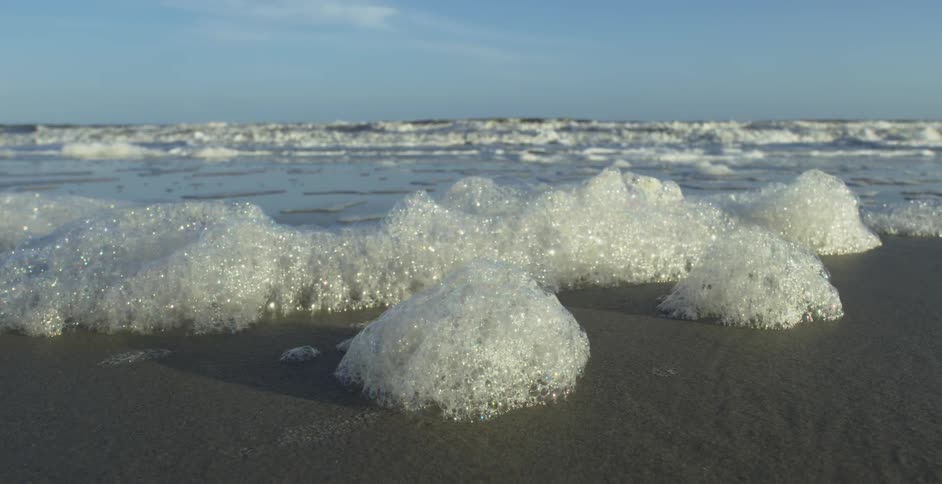 A low-angle close-up shot of sea foam resting on a sandy beach as ocean waves gently roll in the background
