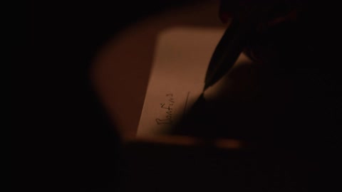 A close-up shot of a hand writing in a diary with a pen in a dark, candlelit room