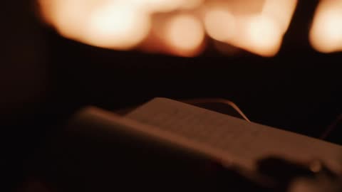 A person's hand writes in a journal with a pen, silhouetted by the warm light of a crackling fire