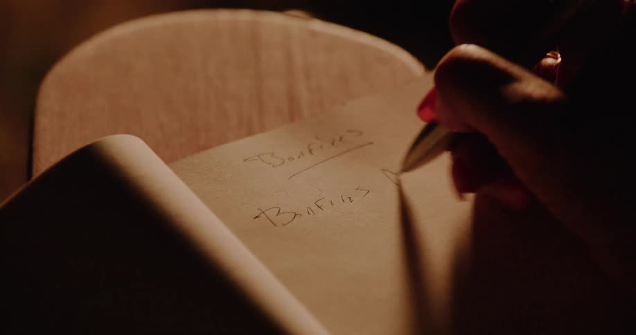Close up of a woman's hand with red nails writing song lyrics about bonfires in a notebook by warm light