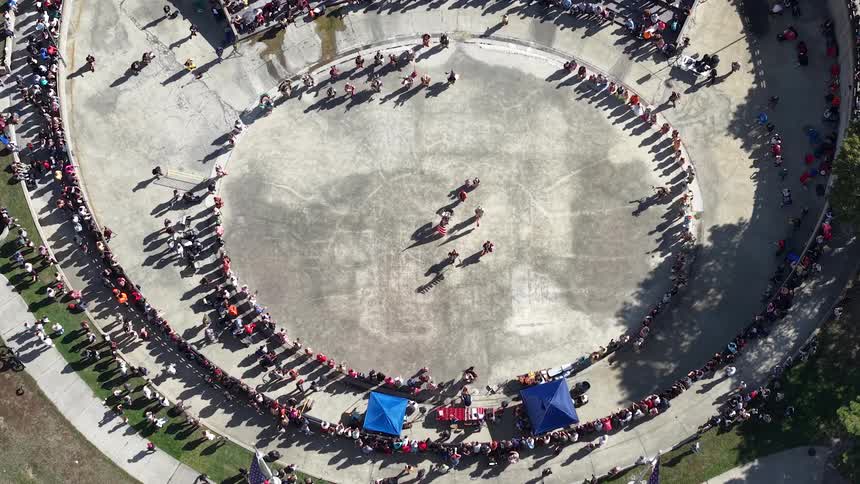 An aerial top-down perspective captures a massive crowd forming a circle around performers during a cultural event in an open-air arena
