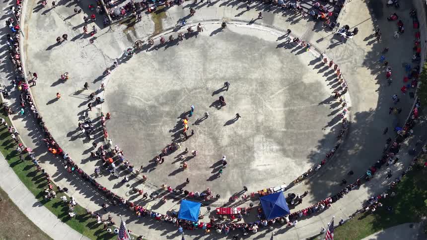 An aerial top-down view shows a community parade moving through a circular plaza as crowds of spectators watch