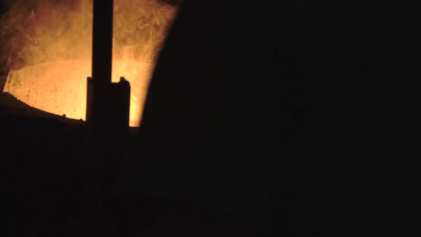 A large vat of molten metal glows intensely and emits smoke in a dark industrial factory