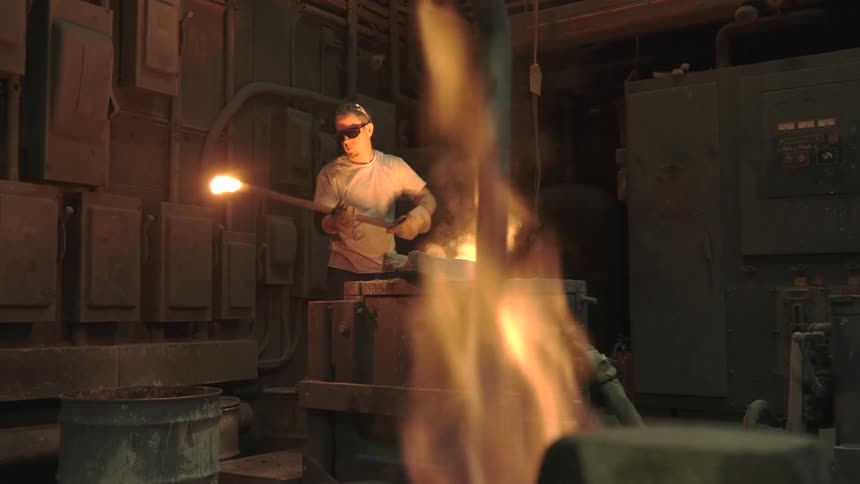 A foundry worker in safety gear uses a long rod to work with molten metal in an industrial furnace