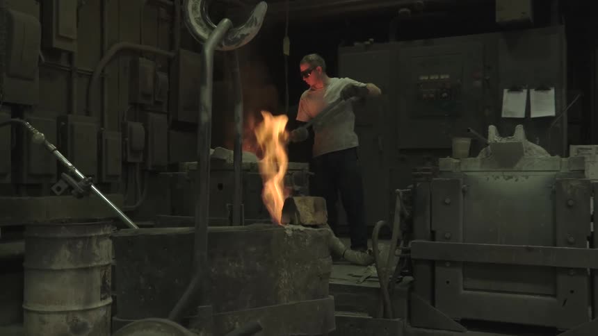 A foundry worker wearing safety glasses uses a long rod to stir molten metal in a fiery crucible inside a dark industrial factory