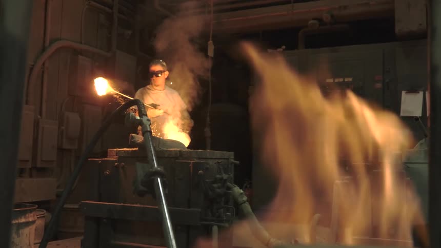A steelworker in protective gear carefully handles glowing molten metal in a dark industrial foundry