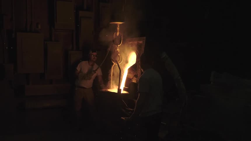 In a dark factory, two men work together to pour glowing molten metal from a large suspended crucible