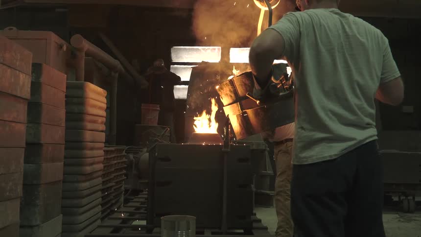 Factory workers in a dark foundry pour fiery molten metal from a crucible into an industrial mold in slow motion