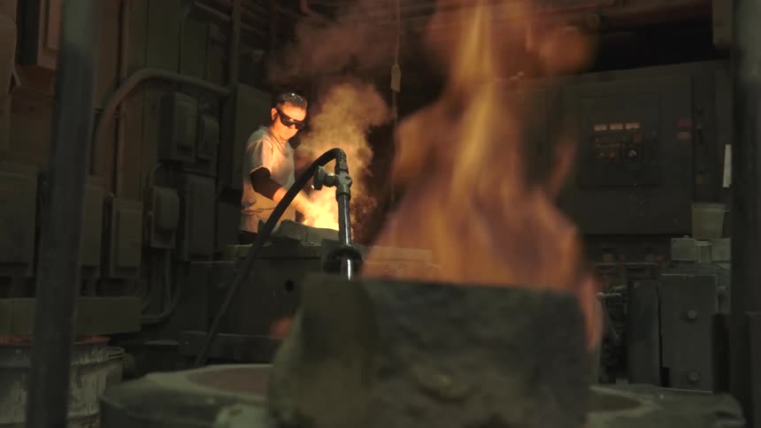 A worker in a hot, dark foundry tends to a furnace of molten metal with large flames burning in the foreground