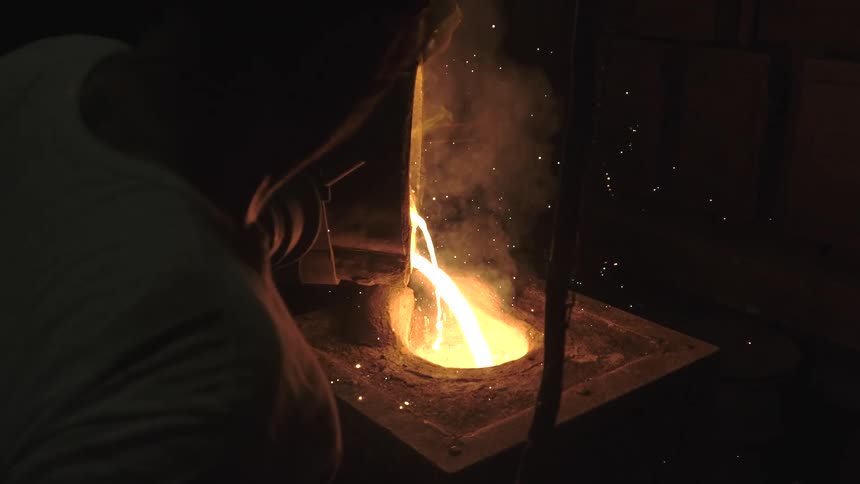 A foundry worker carefully pours glowing molten metal from a crucible into a casting mold in a dark factory, with sparks flying in slow motion