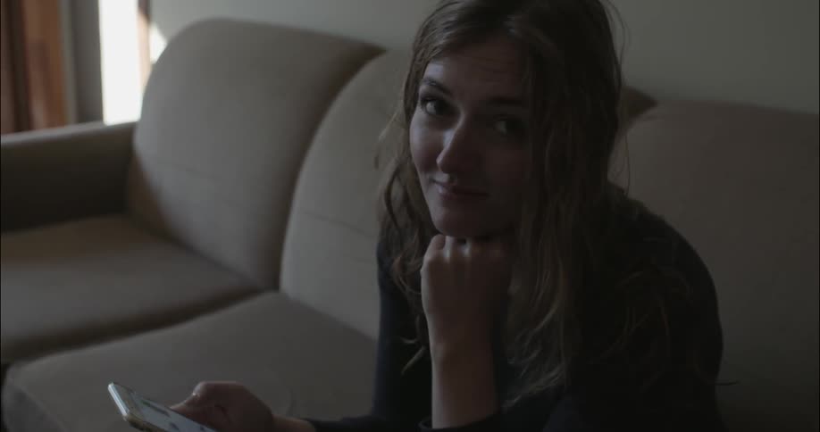 A woman sitting on a couch holding her phones and smiling at the camera