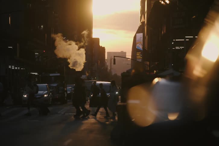 People walk in slow motion across a busy New York City street at golden hour as steam rises from a manhole