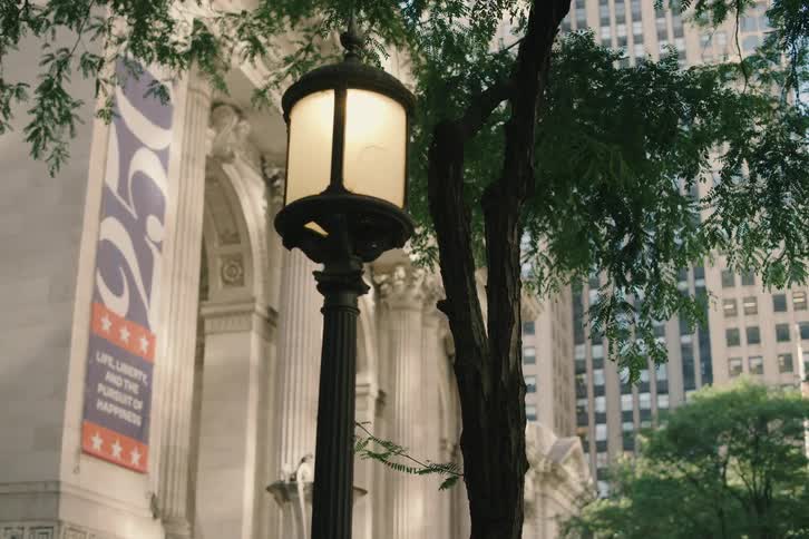 A vintage lamppost stands among lush green trees outside the New York Public Library as the camera slowly reveals the building's classic architecture