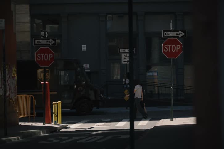 A static shot from a shadowed alley captures pedestrians crossing a sun-drenched urban street with prominent stop signs