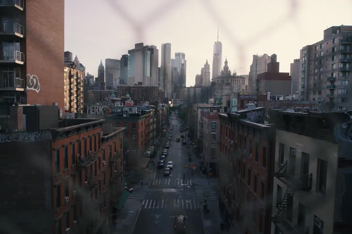 A gritty New York City street leads towards the downtown Manhattan skyline, viewed through a chain-link fence at dusk in a slow-moving shot