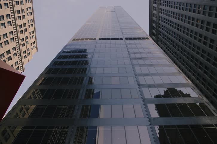 A slow, low-angle tilt-down shot reveals the immense scale of a modern glass skyscraper in a downtown city environment