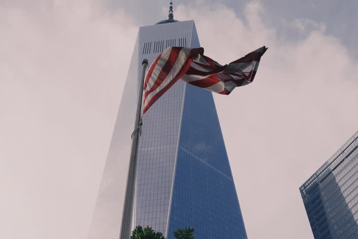 An American flag waves in slow motion in front of the One World Trade Center in New York City against an overcast sky