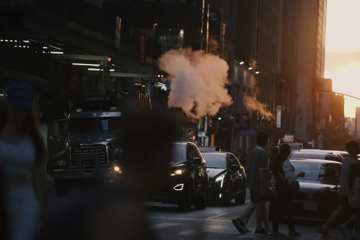 Slow motion captures a crowded New York City street crossing at sunset, with steam from a manhole cover backlit by the golden light