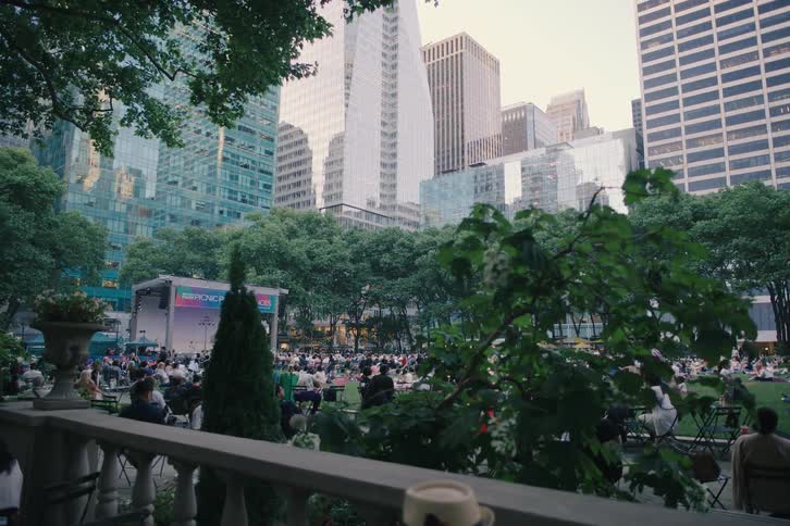 A slow-moving shot captures the serene atmosphere of an evening picnic performance in a bustling city park, surrounded by impressive skyscrapers