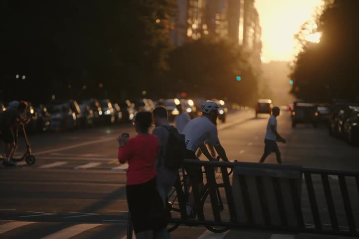 A cyclist waits at a city crosswalk during a beautiful golden hour sunset as traffic and pedestrians pass by in slow motion