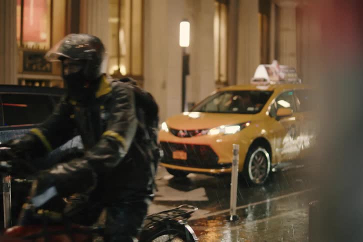 A food delivery cyclist navigates a busy city street in the pouring rain at night, with a yellow taxi cab in the background