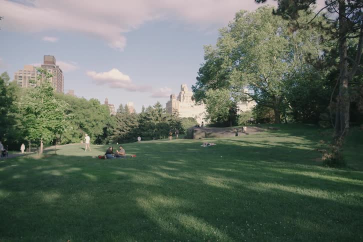 A slow pan reveals people relaxing and picnicking on a grassy hill in Central Park on a sunny day with New York City buildings in the background
