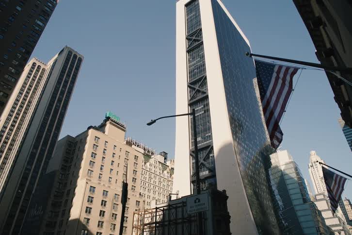 A slow pull-back from a low-angle shot reveals towering modern skyscrapers against a clear blue sky, with an American flag coming into view