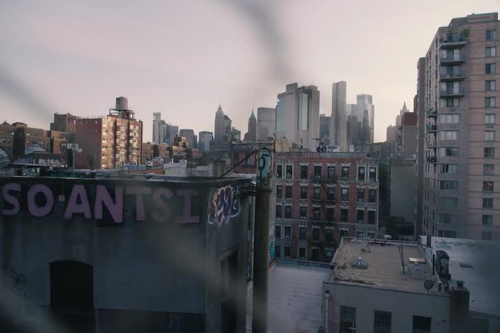 A gritty view of the New York City skyline from a rooftop, looking past graffiti-covered buildings through a chain-link fence