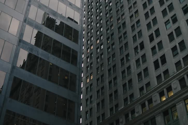 A low-angle view slowly tilts up on a modern glass skyscraper reflecting a classic building as office lights begin to turn on at dusk