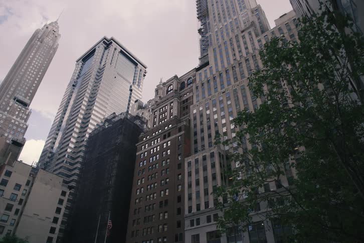 A slow reveal from behind a building unveils a low-angle view of various towering skyscrapers in downtown Manhattan on an overcast day