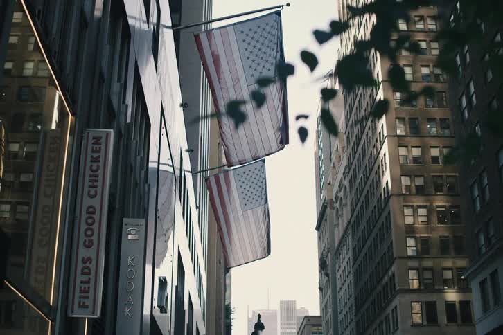 A cinematic shot reveals two American flags hanging between tall city skyscrapers as the camera slowly moves forward