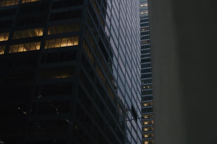 A slow pan reveals the contrasting facades of modern city skyscrapers at dusk, with warm lights glowing from within