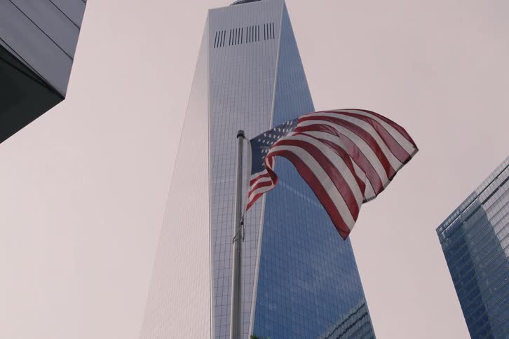 The American flag waves in slow motion with the One World Trade Center skyscraper towering in the background