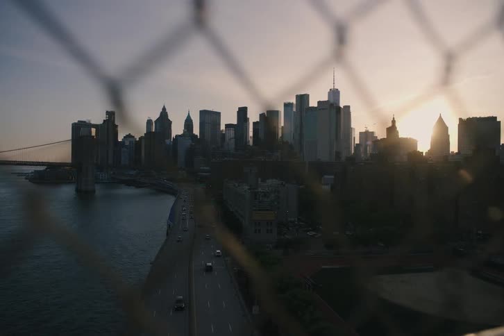 A slow-motion view of the Lower Manhattan skyline and FDR Drive at sunset, framed through a chain-link fence on a bridge