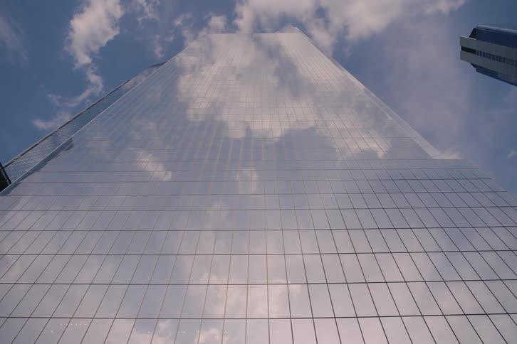 A low-angle slow pull-back shot captures the reflection of clouds moving across the glass facade of a modern skyscraper reaching into the blue sky