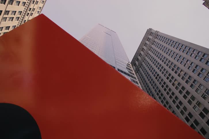 A slow, low-angle tilt down from imposing city skyscrapers reveals a large, modern red sculpture on the street below