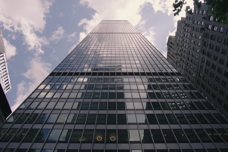 A slow, dramatic low-angle dolly shot looking up at a modern glass skyscraper, with clouds reflected in its facade and a tree moving into the foreground