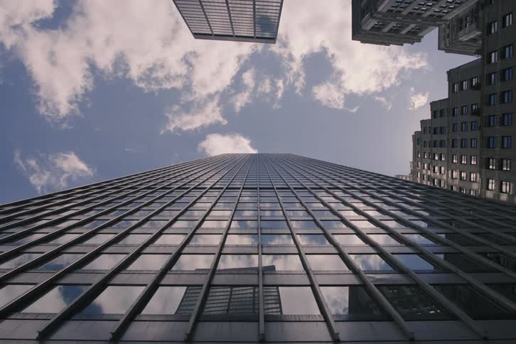 Dynamic time-lapse from a low angle shows clouds passing over a towering glass skyscraper, their reflections moving across the windows