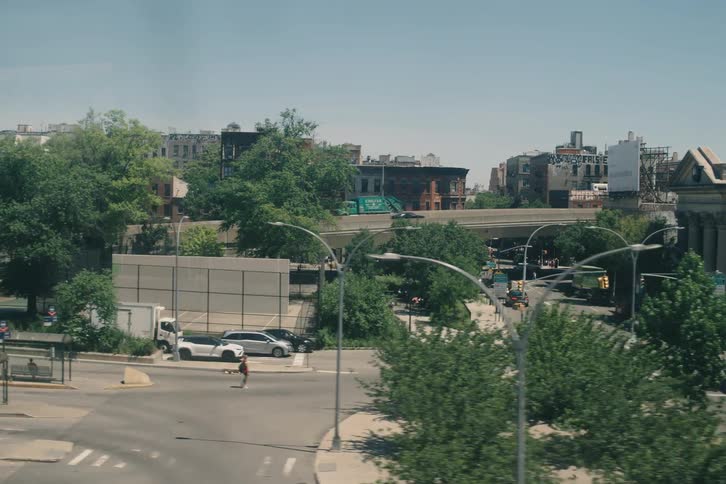 A slow-motion tracking shot from a moving vehicle passes by graffiti-covered rooftops and trees, revealing a sunny urban street scene