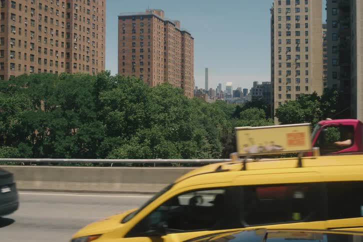 Slow-motion tracking shot of bustling highway traffic in New York City on a sunny day, with large apartment buildings and trees in the background