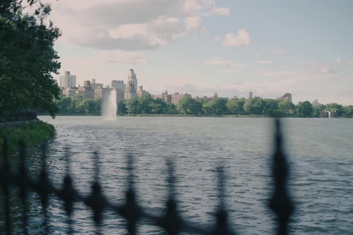 Tracking shot past a black iron fence reveals the Jacqueline Kennedy Onassis Reservoir with a fountain and the Upper West Side skyline in the background