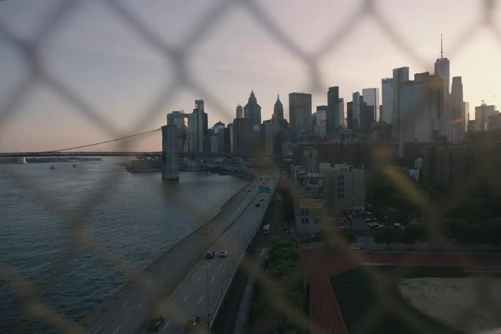 The Lower Manhattan skyline and Brooklyn Bridge are viewed through a chain-link fence at sunset as traffic flows on the highway below
