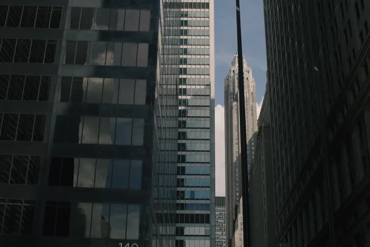 A slow dolly shot moves through a shadowed city street, looking up at towering skyscrapers and their reflections