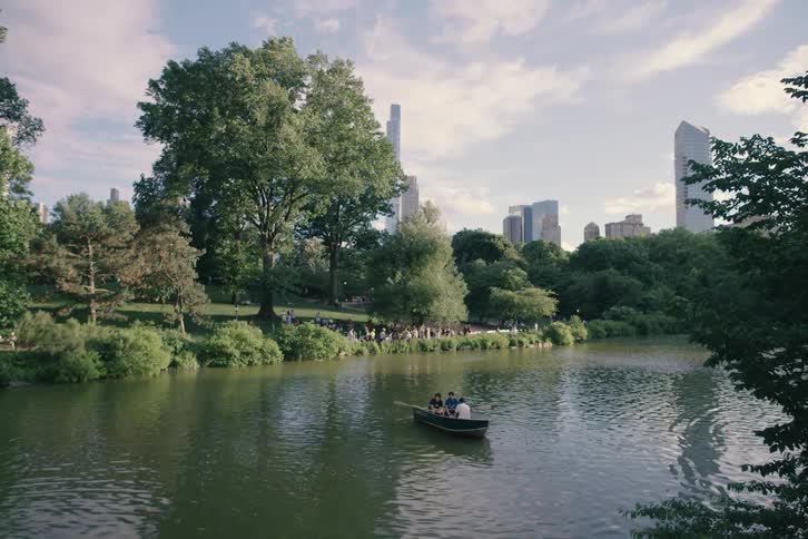 A beautiful slow reveal shows people rowing a boat on the lake in Central Park, with the iconic New York City skyline in the distance