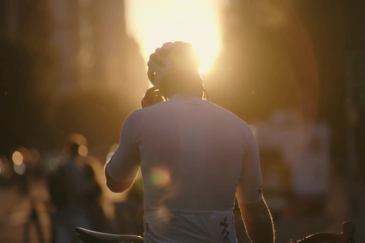 A cyclist stands with his bike on a city street, silhouetted by the golden glow of the setting sun