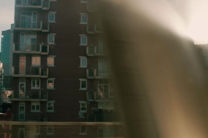 A cinematic view from a moving train reveals the Lower Manhattan skyline and gritty urban buildings bathed in the warm glow and flare of the setting sun