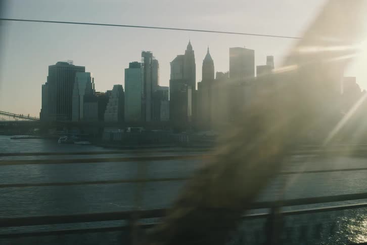 A silhouetted view of the Lower Manhattan skyline and Brooklyn Bridge from a moving vehicle as the morning sun creates a lens flare